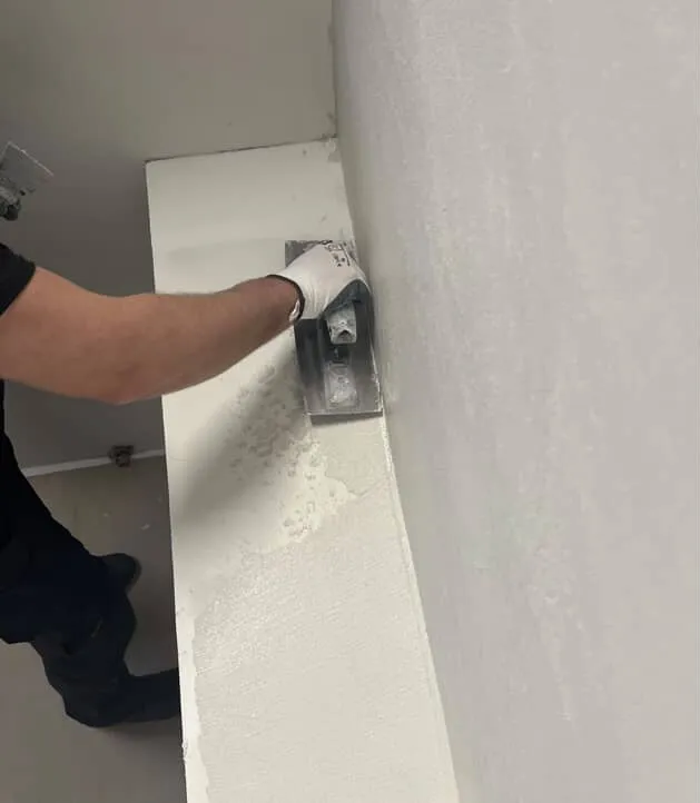 Työntekijän hansikoitu käsi hiertää mikrosementtiä seinän reunaan teräslastalla.