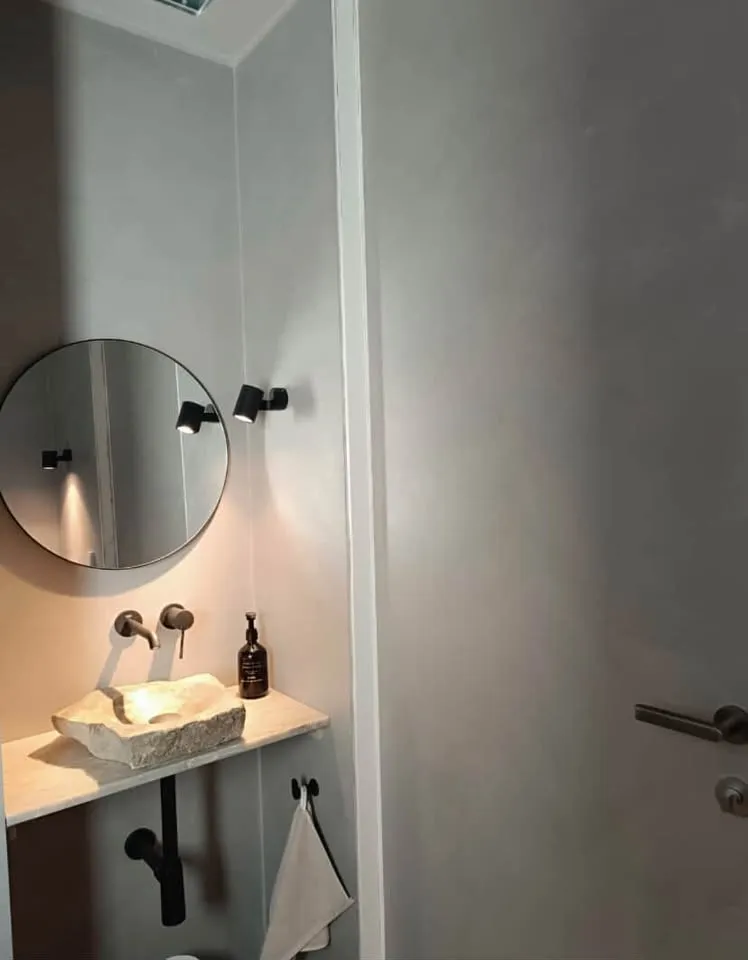 Pieni WC-tila, jossa kivimalja-allas, pyöreä peili ja mikrosementtiseinä.